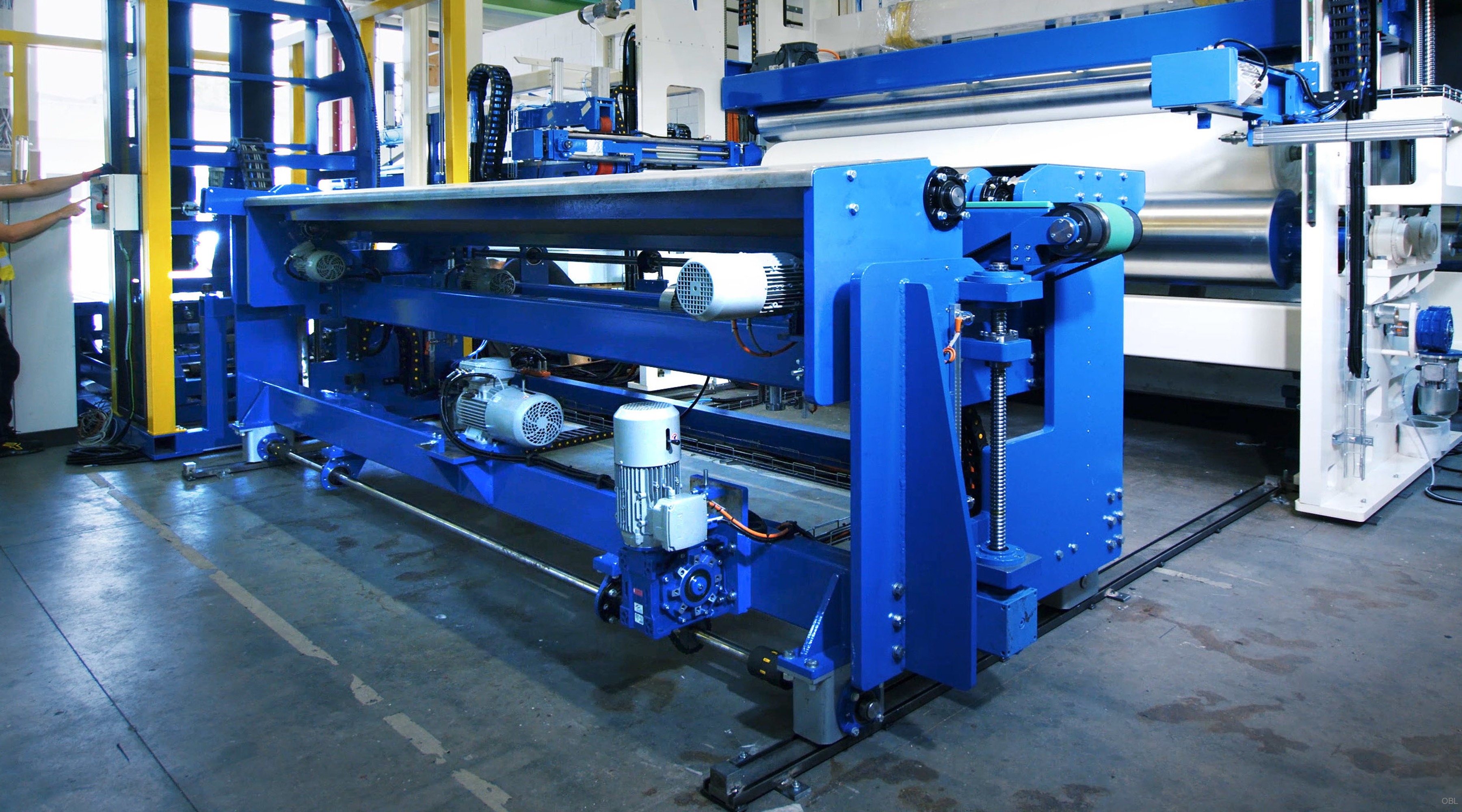 The photograph displays an industrial setting where construction machinery operates within a large facility. A prominent piece of blue machinery takes center stage; it appears to be part of a complex system, possibly involved in textile or material handling given its structure and components such as rollers, gears, and support frames. The surrounding area suggests that this environment is designed for heavy-duty work with other similar equipment visible in the background.