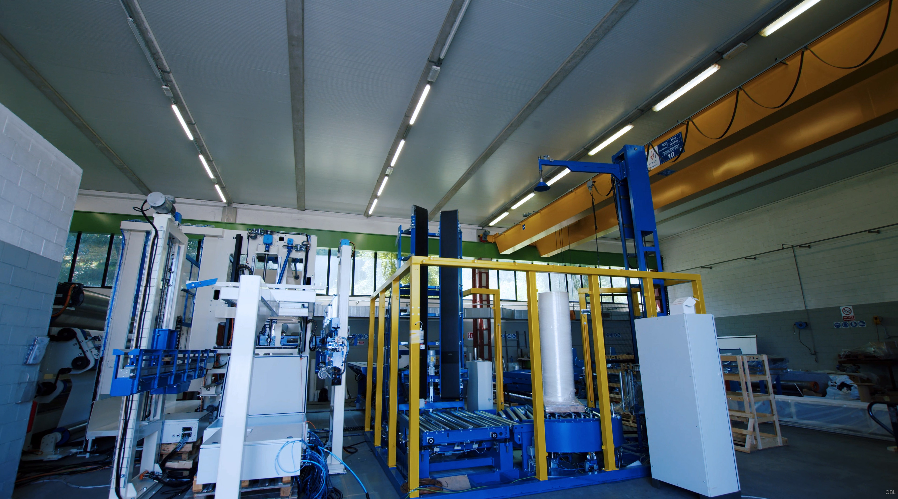 La foto cattura un ambiente industriale con vari componenti meccanici in bella mostra. Una caratteristica centrale dello spazio è una grande struttura blu e gialla a forma di cornice, probabilmente parte dei macchinari o infrastruttura a supporto del progetto macchina ewrapper. Intorno a questo componente principale ci sono altre macchine e attrezzature dipinte in diverse tonalità di blu, bianco e grigio, che ne indicano la diversità funzioni all'interno dell'installazione. Il soffitto della stanza ospita più luci fluorescenti che illuminano lo spazio di lavoro sottostante. Le pareti sembrano essere costruite con materiali robusti come il cemento, fornendo un ambiente robusto per le attività industriali. Sullo sfondo si possono vedere ulteriori macchinari o parti impilati contro il muro su scaffali, suggerendo un ambiente organizzato e organizzato struttura ben mantenuta dedicata alle attività di produzione o assemblaggio. Nel complesso, questa immagine offre uno sguardo sugli aspetti tecnici della costruzione di macchine per l'imballaggio avanzate, mostrandone sia la complessità le attrezzature coinvolte e l'ambiente strutturato in cui viene sviluppato.